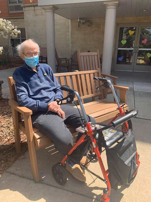 An elderly man wearing a blue face mask, blue sweatshirt, gray pants, and brown slippers is sitting on a wooden bench outdoors. In front of him is a red walker with a black seat and storage bag. Behind him is a building entrance with glass doors decorated with colorful heart-shaped stickers.