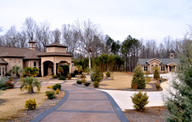 View of a senior living facility with a paved walkway leading to two beige buildings surrounded by landscaped bushes and trees, with a backdrop of leafless trees under a clear sky.