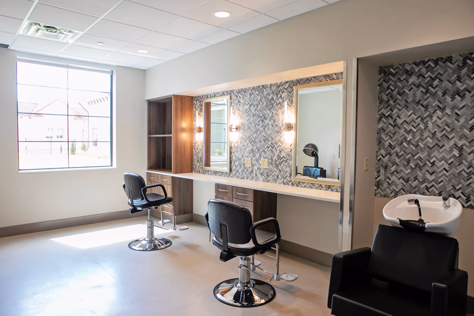 A bright salon area with two black styling chairs in front of a long counter with two mirrors and wall-mounted lights. There is a black shampoo chair with a white sink on the right side. The walls have a patterned gray and white tile design, and a large window lets in natural light.