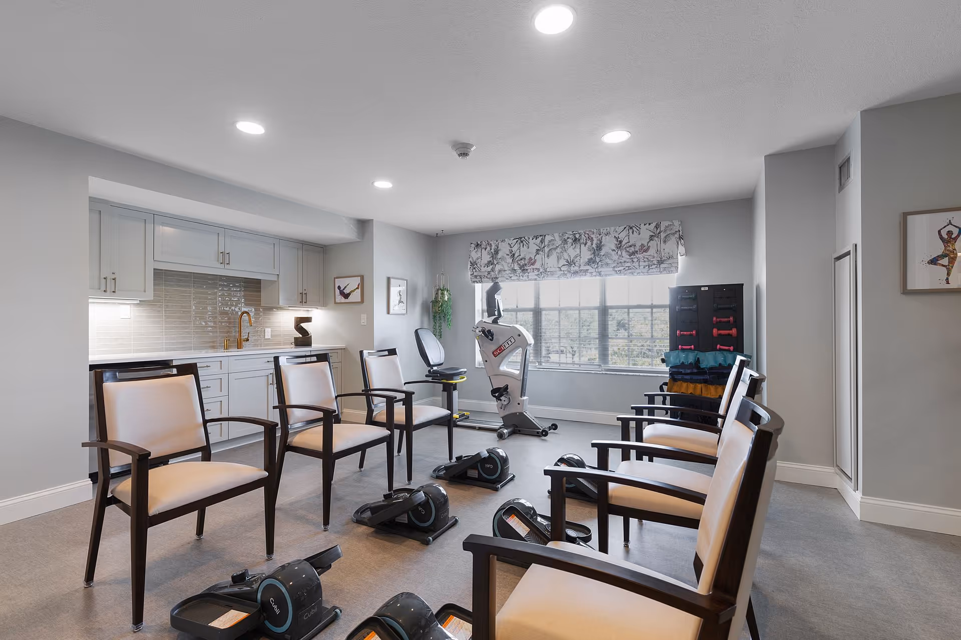 A bright exercise room with six beige cushioned chairs arranged in a circle. In the center are several compact exercise machines. The room has light gray walls, a large window with floral valance, and a stationary exercise bike near the window. There is a small kitchenette with white cabinets and a tiled backsplash on the left side, and a rack with weights and exercise mats near the window.