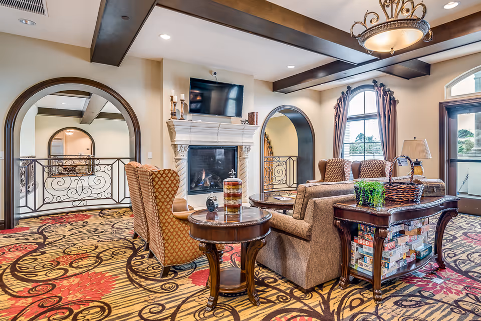 Cozy common room with sofas and armchairs arranged around a fireplace and wall-mounted TV, ornate rug, chandelier and arched doorways.