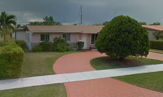Single-story residential building with a pink exterior and a brown roof, surrounded by a lawn with bushes and a large tree near a curved red brick driveway leading to the entrance.