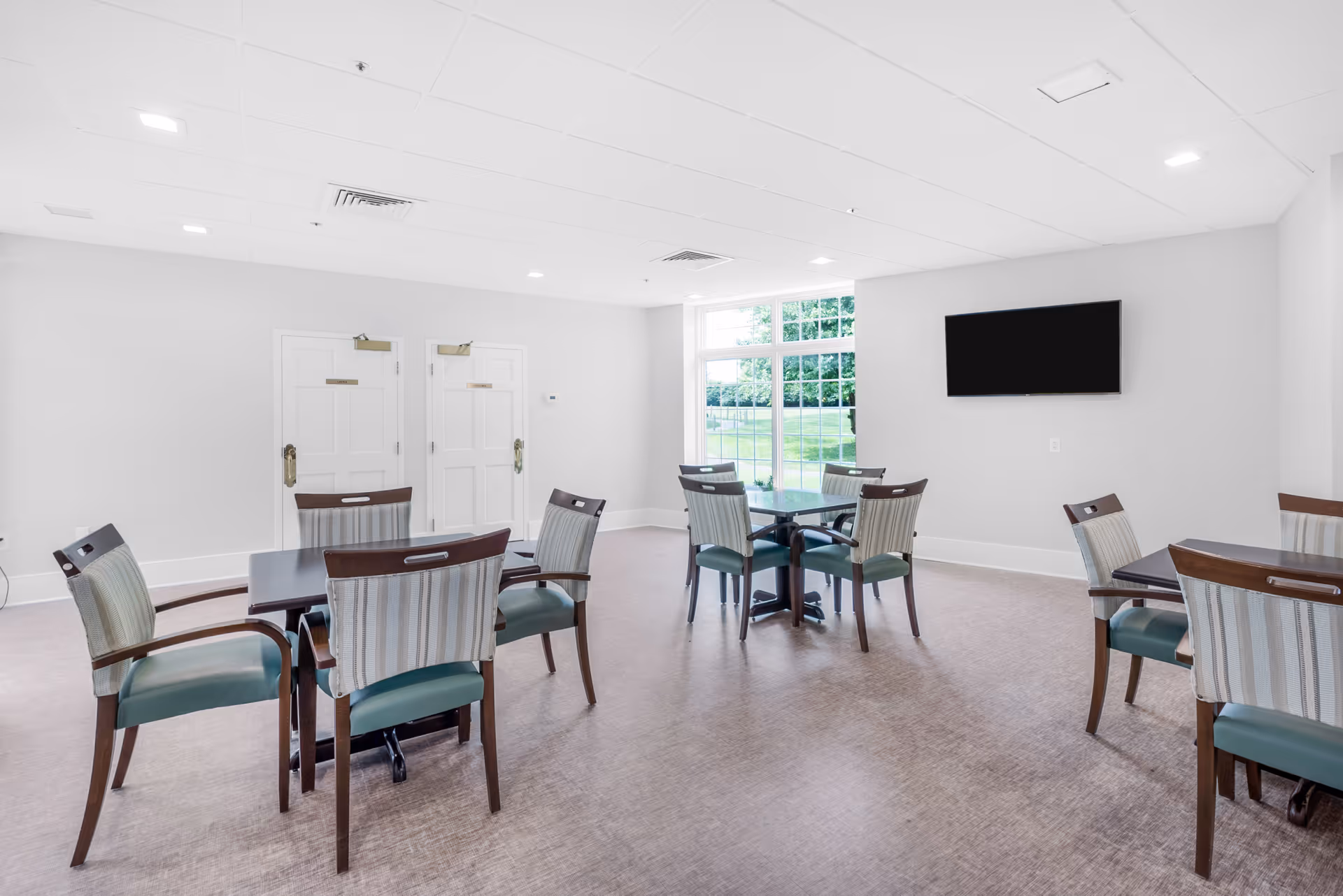 A bright and clean dining area in a senior living facility with several square tables surrounded by wooden chairs with green cushions and striped backs. There is a large window letting in natural light and a flat-screen TV mounted on the wall. Two white doors are visible on the far wall.