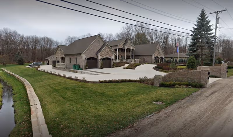 A large, single-story brick building with multiple peaked roofs and a spacious driveway surrounded by well-maintained green lawns and trees in the background under a cloudy sky.