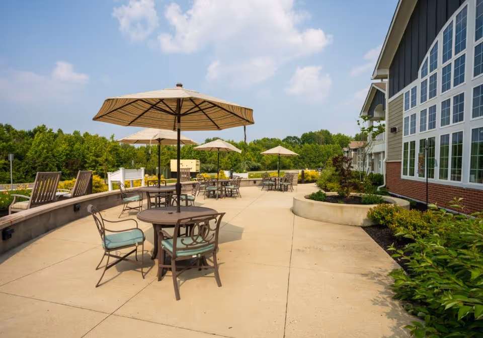 Outdoor patio area with round tables and chairs under large striped umbrellas, surrounded by greenery and adjacent to a building with large windows under a partly cloudy sky.