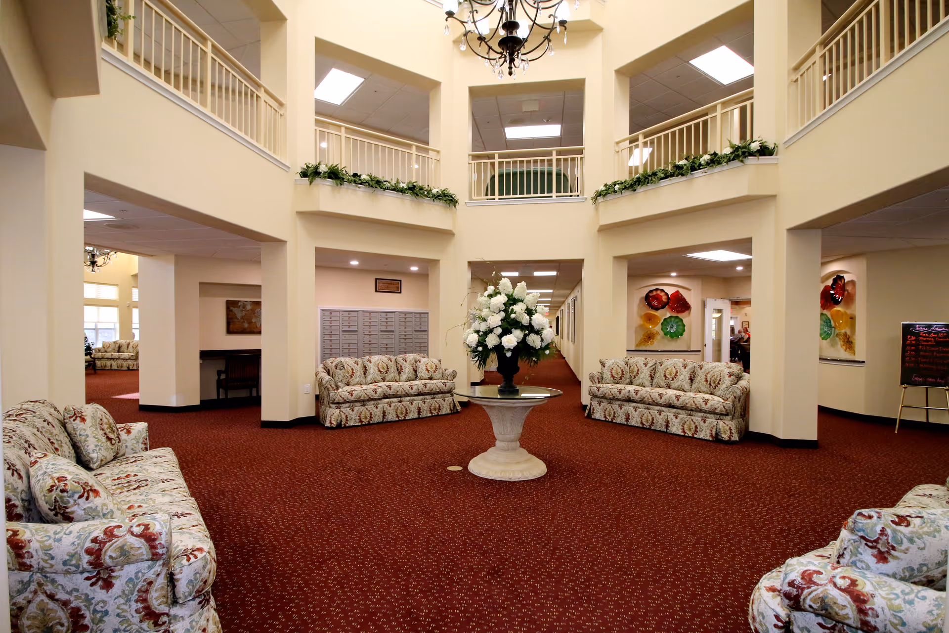 Spacious and well-lit common area in a senior living facility with patterned sofas arranged around a central round table holding a large floral arrangement. The room features high ceilings with a chandelier, beige walls, red carpet, and upper-level balconies with railings. Decorative plants and colorful wall art are visible in the background.