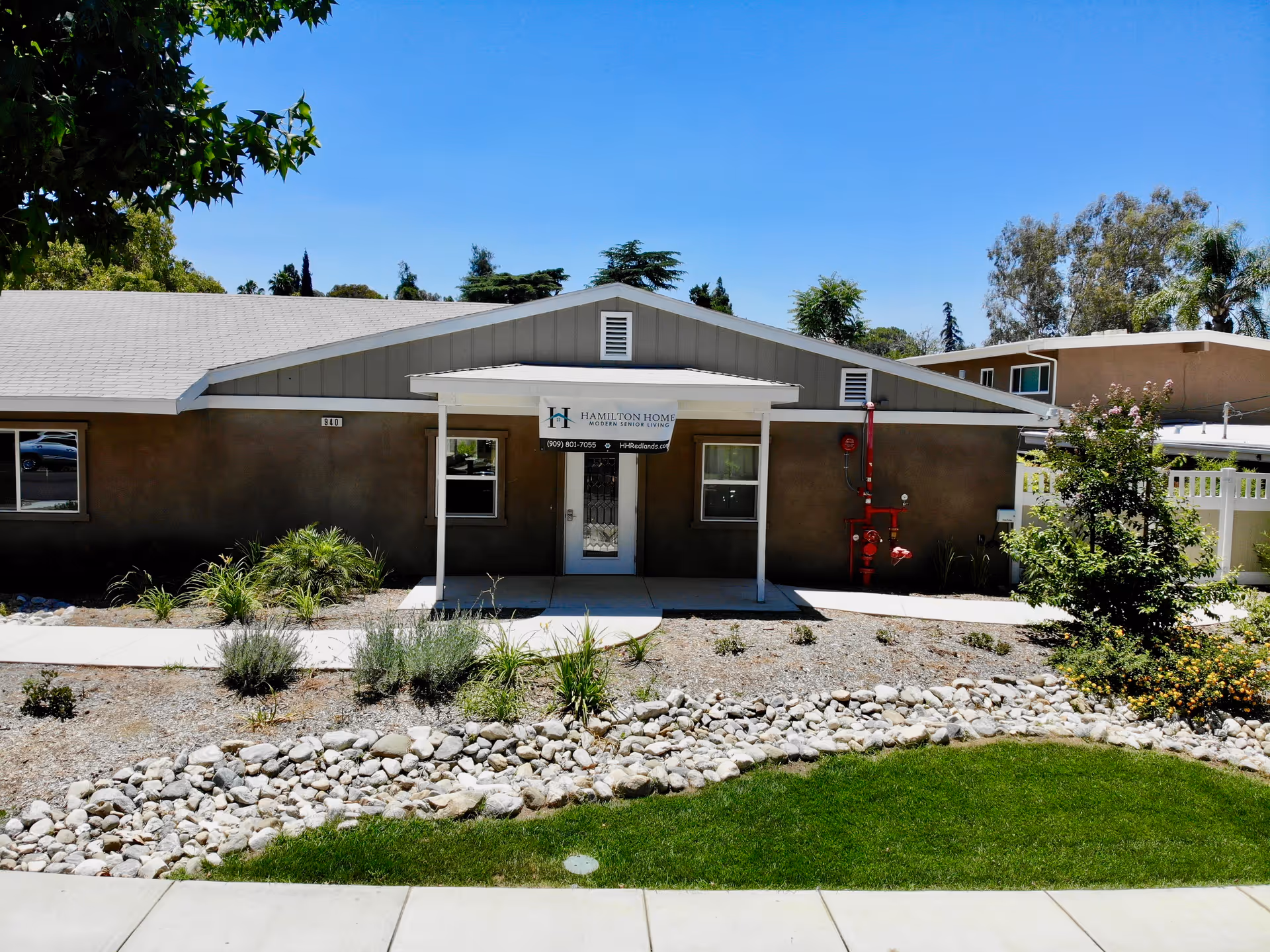Exterior view of a single-story building with a gray roof and brown walls, featuring a small covered entrance with a sign that reads 'Hamilton Home Modern Senior Living'. The building is surrounded by a landscaped area with rocks, plants, and a green lawn under a clear blue sky.