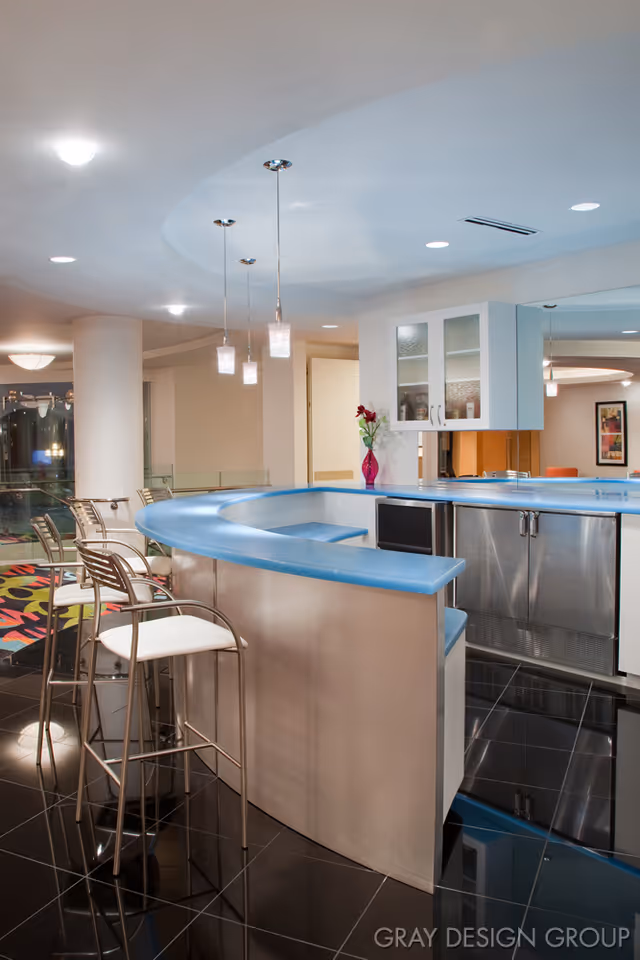 Modern indoor bar area with a curved blue countertop, metal bar stools with white cushions, pendant lights hanging from the ceiling, and a small vase with flowers on the counter. The floor is shiny black tile, and there are cabinets and a mini fridge behind the bar.