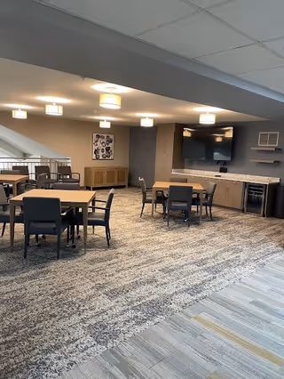 A spacious common area with multiple square tables and chairs arranged for seating. The room features a carpeted floor with a patterned design, a large flat-screen TV mounted on the wall, and cabinetry with a countertop below the TV. Ceiling lights provide illumination, and there is a decorative framed artwork on the far wall.