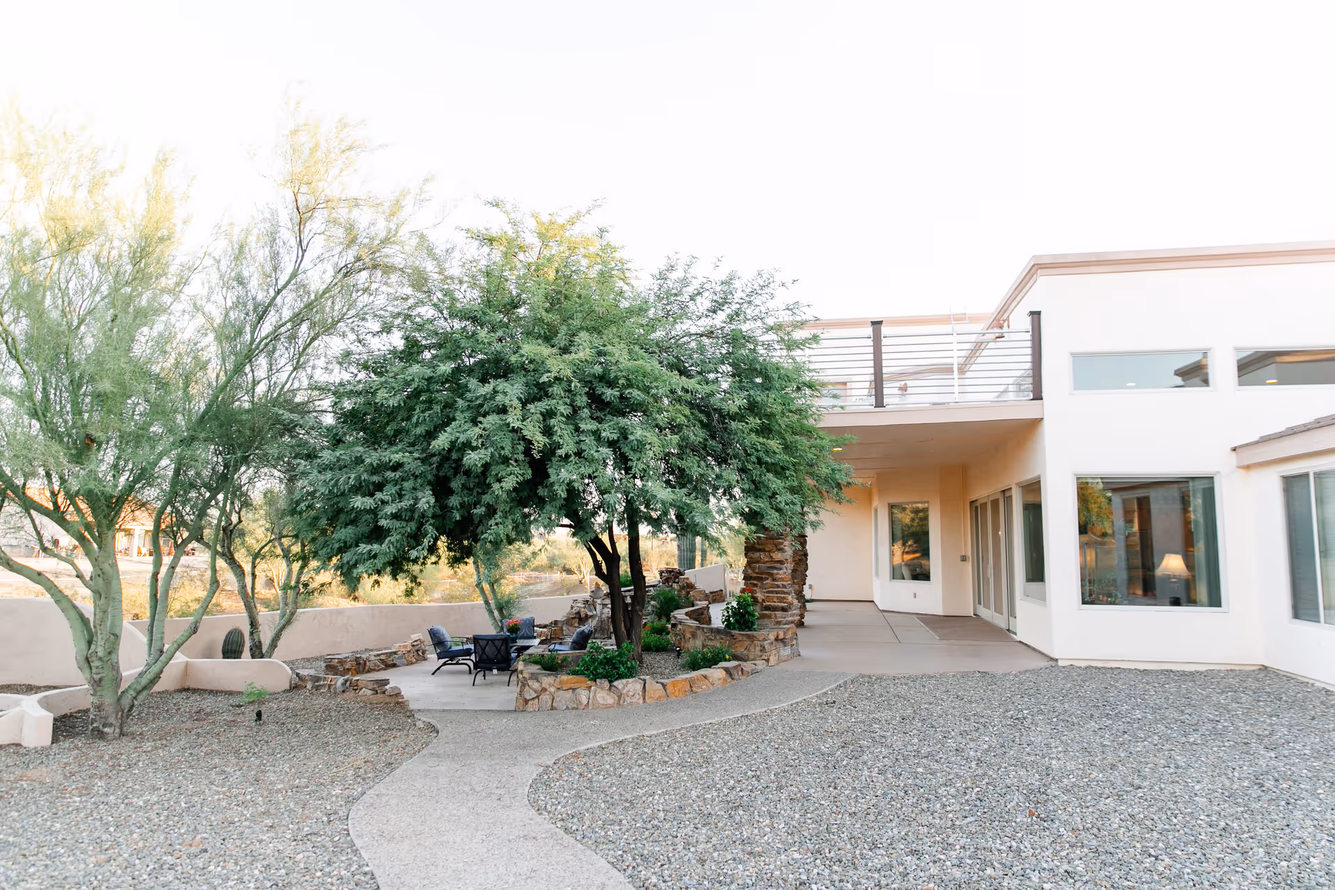 Outdoor patio area at Carefree Assisted Living featuring a curved concrete pathway, gravel ground cover, a large leafy tree surrounded by a stone planter, several chairs arranged around a small table, and a white building with large windows and a balcony.