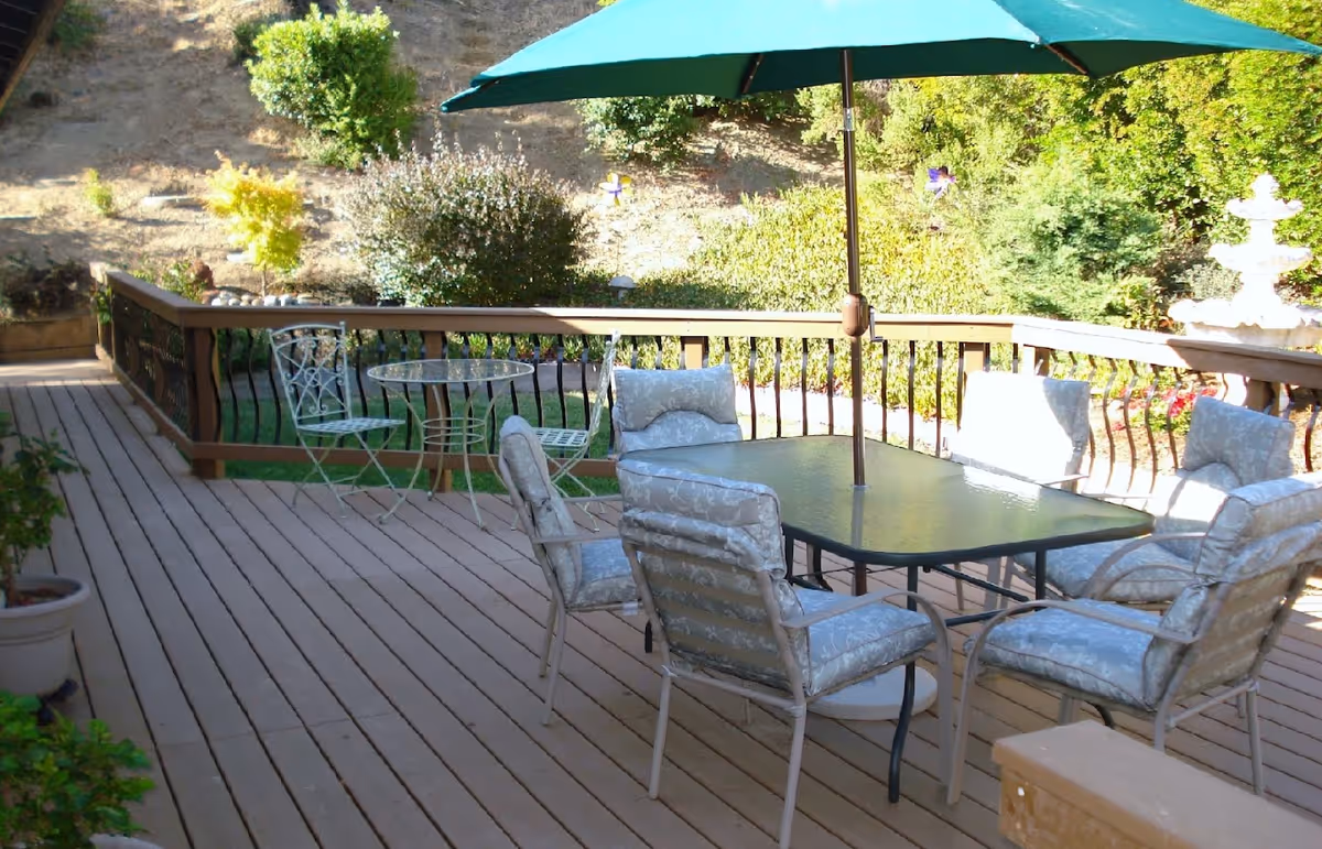 Outdoor patio area with a glass table and six cushioned chairs under a large green umbrella. There is also a small round metal table with two matching chairs. The patio is surrounded by a wooden railing and overlooks a garden with bushes, trees, and a white fountain in the background.