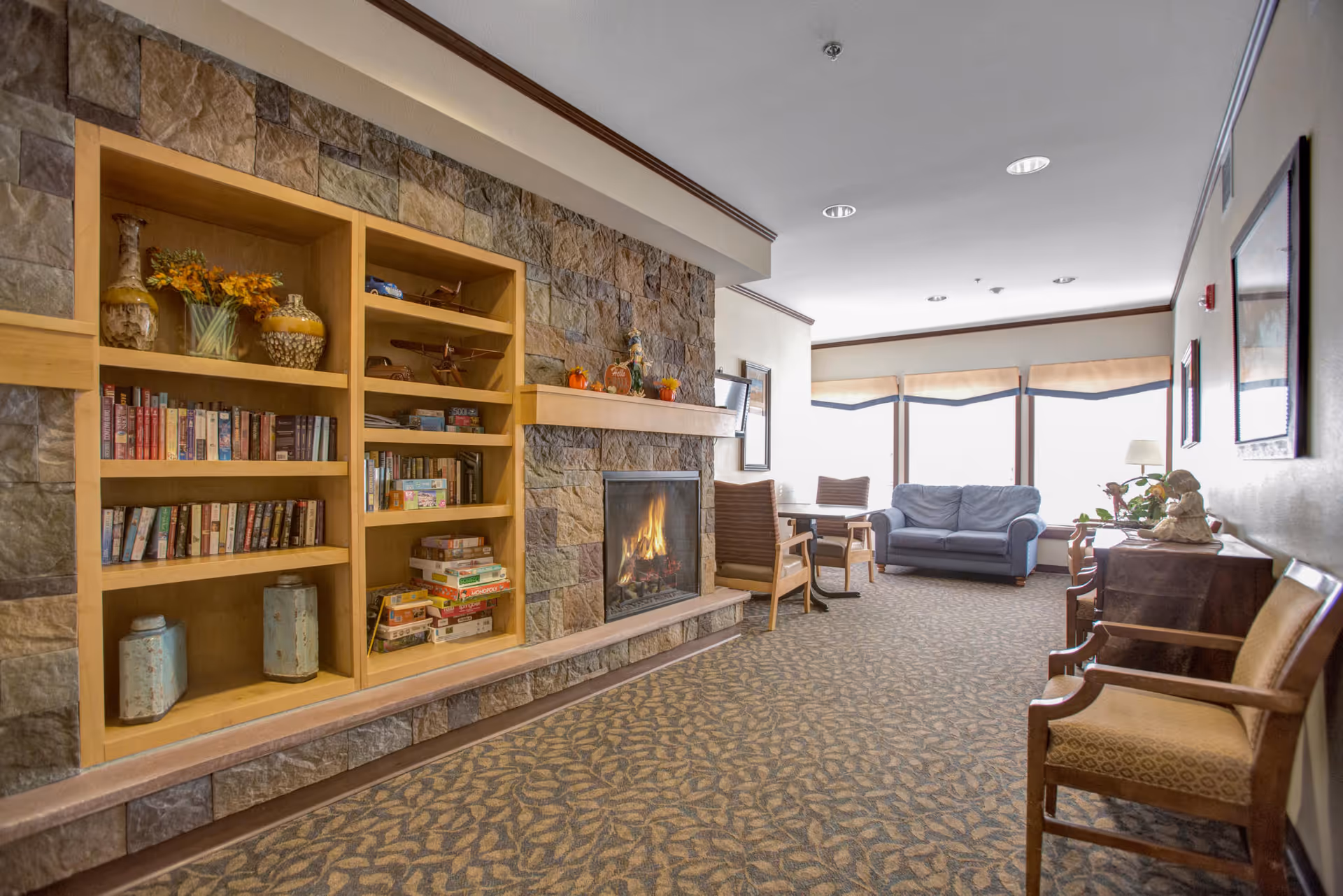 A cozy common lounge with built-in bookshelves and a lit stone fireplace, chairs and a sofa by large windows.