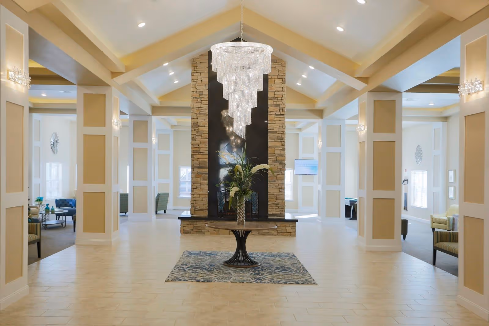 Spacious and elegant interior lobby area with a high vaulted ceiling, a large modern chandelier, a stone fireplace in the center, and a round table with a floral arrangement on a patterned rug. The space is well-lit with wall sconces and natural light from windows, and seating areas with chairs are visible on both sides.