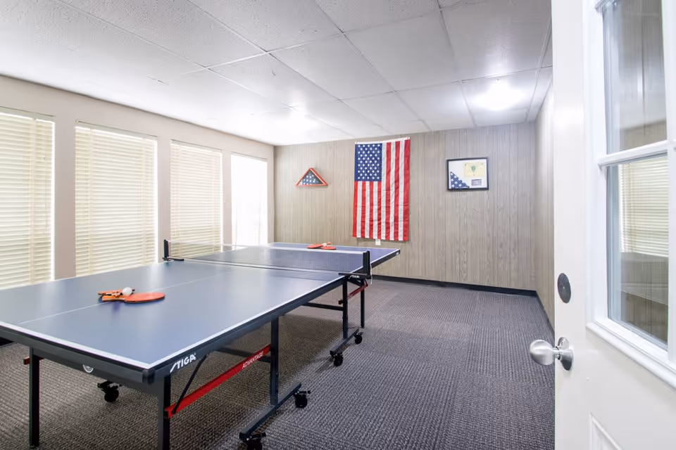 A bright room with a ping pong table in the center, two paddles and a ball on the table. The room has carpeted flooring, wood-paneled walls, and several windows with closed blinds. An American flag and two framed displays with folded flags are mounted on the far wall. A white door with a glass window is partially open on the right side.