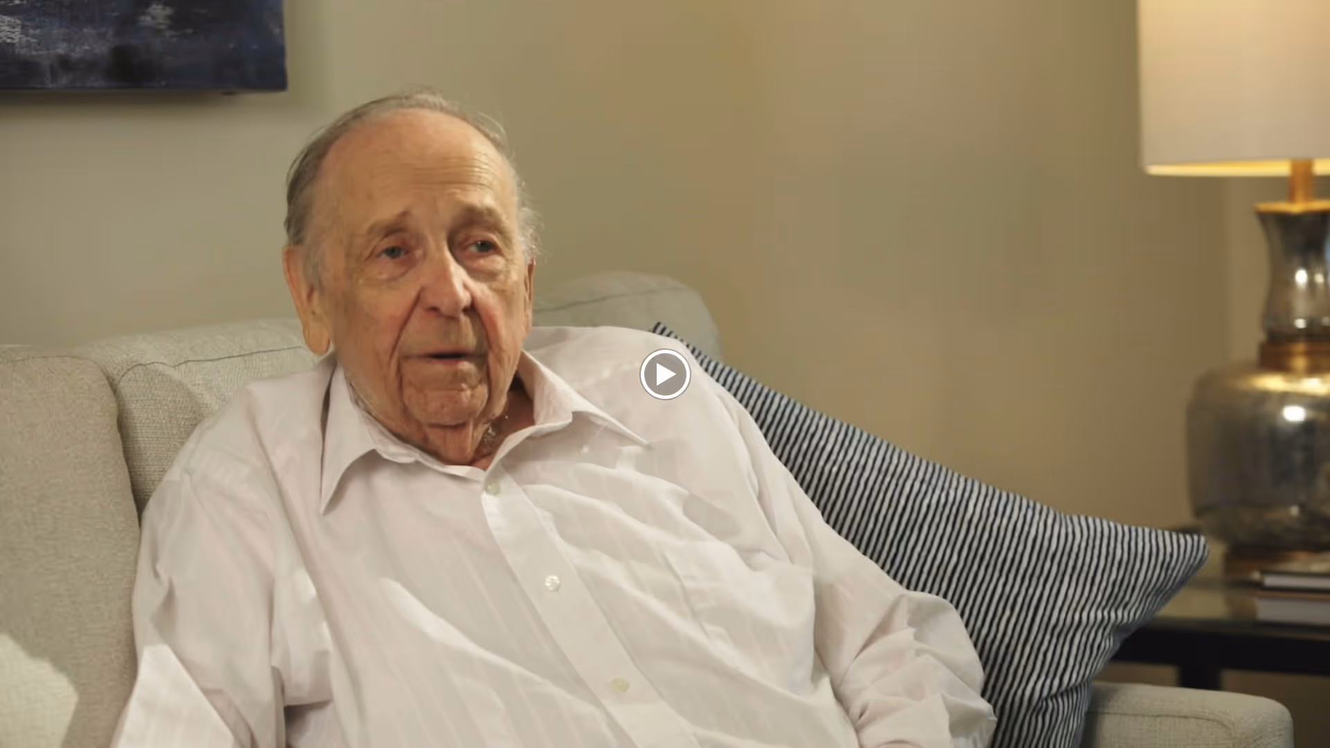 An elderly man wearing a white shirt is sitting on a light gray couch with a striped pillow behind him. There is a lamp with a metallic base and a beige lampshade on a side table next to the couch.