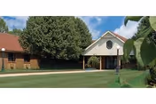 Front view of a senior living facility with brick and white facade, lush lawn, and trees.