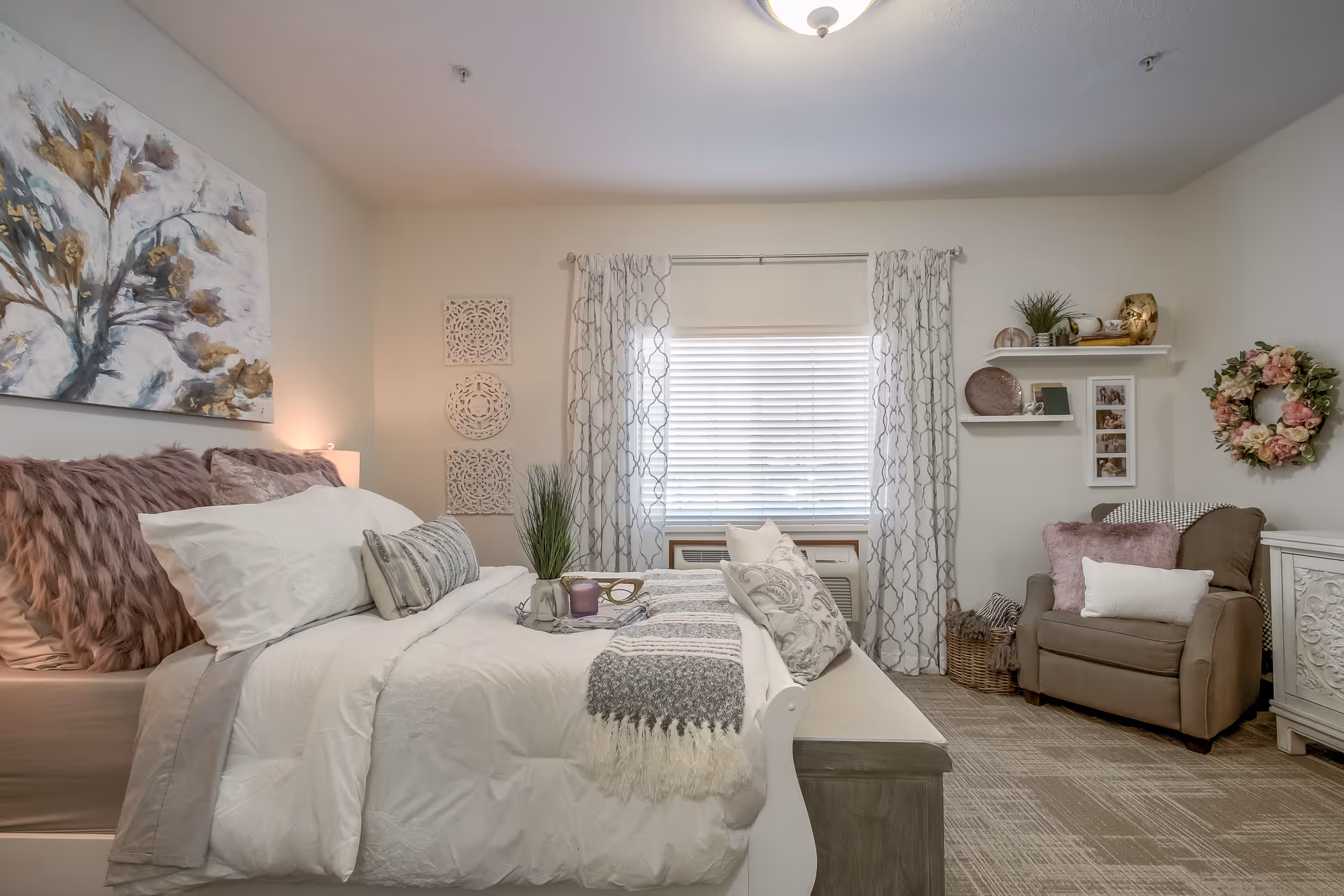 A cozy and well-decorated bedroom in a senior living facility featuring a neatly made bed with white and gray bedding, decorative pillows, and a throw blanket. The room includes a comfortable armchair with cushions, a window with patterned curtains, wall art including a floral painting and decorative panels, a small shelf with plants and framed photos, and a floral wreath on the wall.