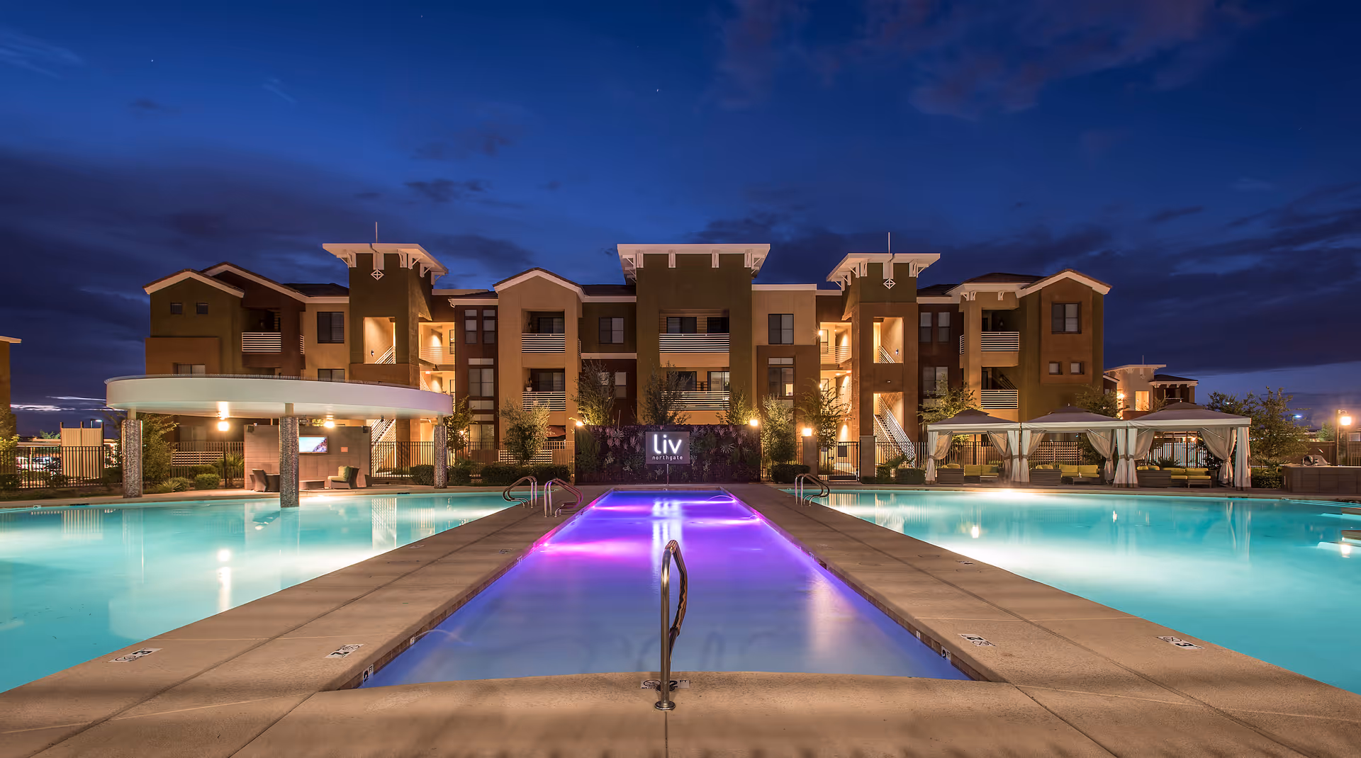 Lit outdoor swimming pool and illuminated central water feature in front of the Liv Northgate apartment building at dusk.