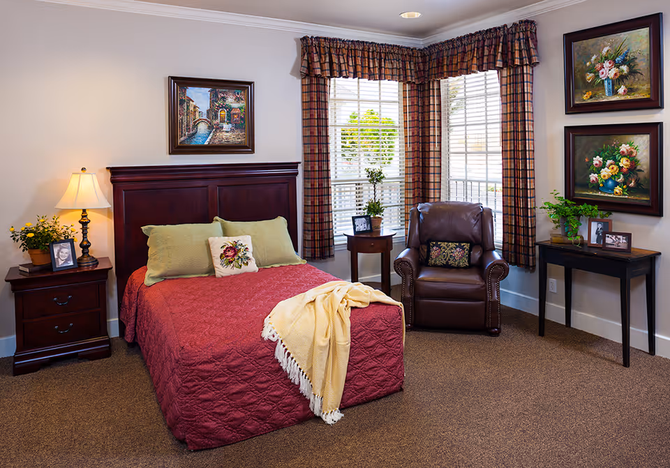 A cozy bedroom with a dark wooden bed frame and a red quilted bedspread, two green pillows and a decorative floral pillow. A yellow throw blanket is draped over the foot of the bed. To the left is a wooden nightstand with a lamp, a potted plant, and a framed photo. A leather armchair with a decorative pillow sits near a corner window with plaid curtains. On the right wall are two framed floral paintings above a small wooden table with a potted plant and two framed photos. The room has beige carpet and white walls.