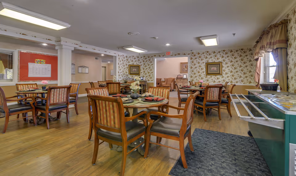 Dining room with round tables set for meals, wooden chairs, a buffet counter, and floral wallpapered walls.