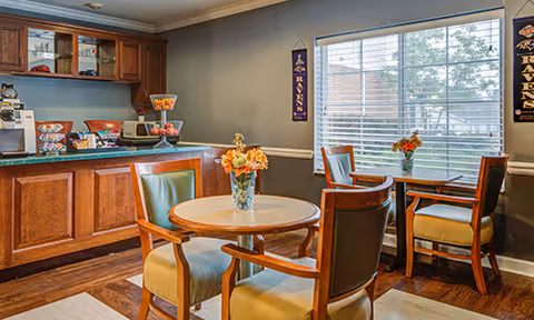 A cozy dining area with wooden tables and chairs featuring green cushions. Each table has a vase with orange flowers. There is a wooden counter with snacks and a coffee machine on the left side. A large window with white blinds lets in natural light, and two Baltimore Ravens banners hang on the gray walls.