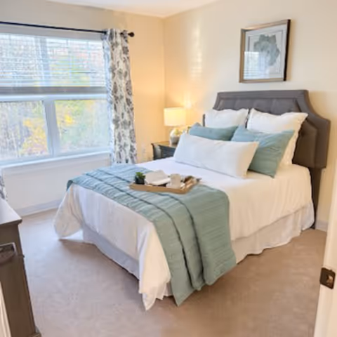 A bright bedroom with a neatly made bed featuring white and light blue bedding, a gray upholstered headboard, a bedside table with a lamp, a framed picture on the wall, and a large window with patterned curtains letting in natural light.