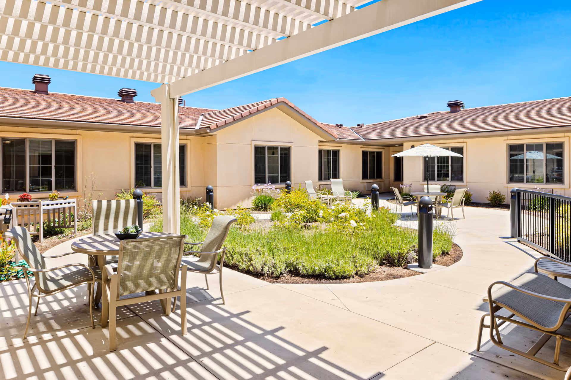 Sunny central courtyard patio with tables, chairs, umbrellas, and landscaped plant beds surrounded by a single-story building.