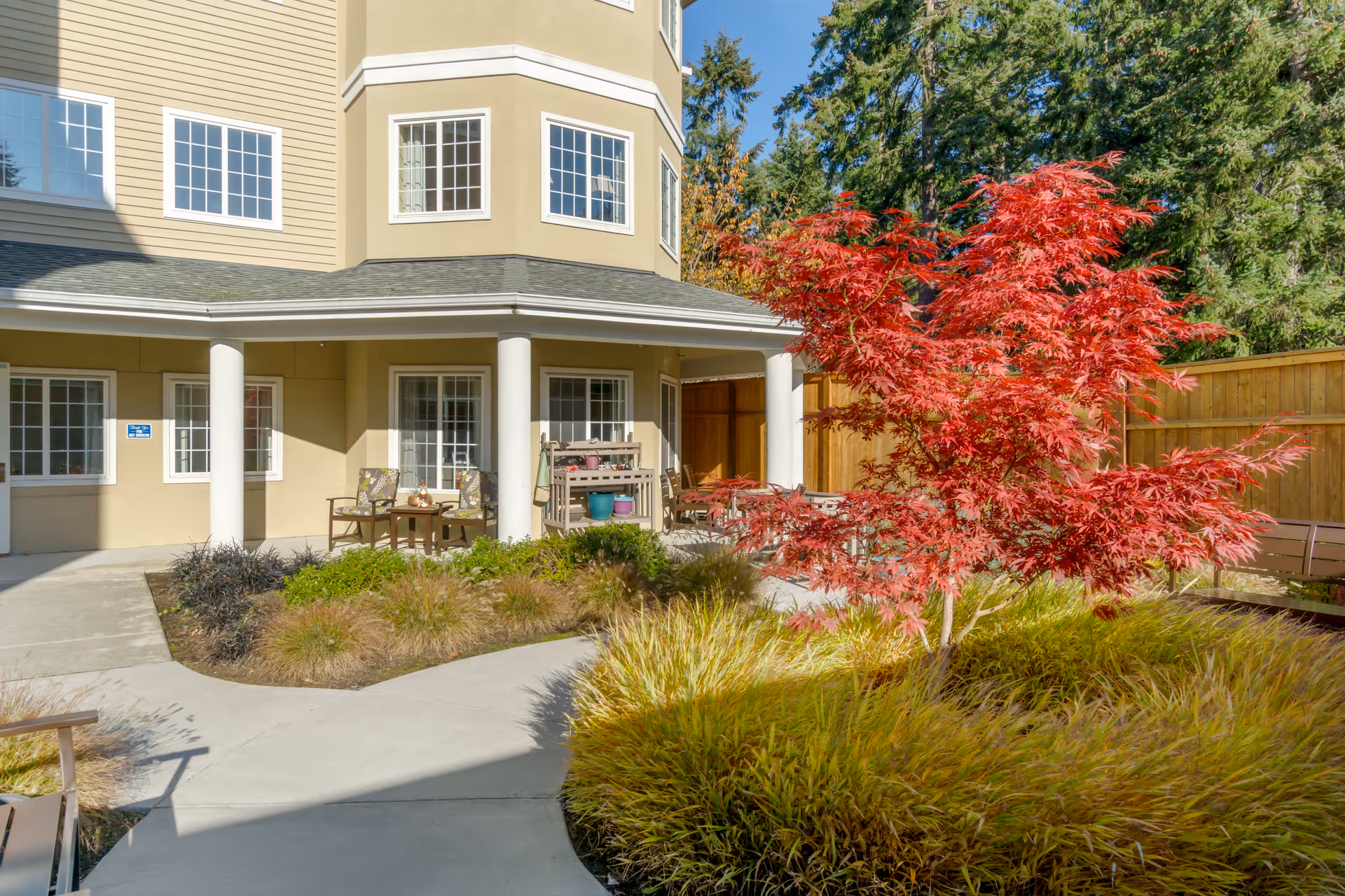 Outdoor patio area of a senior living facility with a beige building featuring multiple windows and white columns. There are chairs and a small table on the patio, surrounded by landscaped greenery including a vibrant red-leafed tree and ornamental grasses. A wooden fence and tall trees are visible in the background under a clear blue sky.
