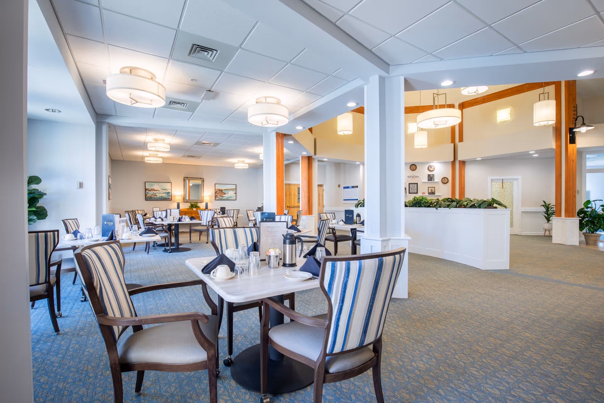 A bright and spacious dining room in a senior living facility with multiple tables and striped upholstered chairs. The tables are set with menus, coffee pots, cups, glasses, and napkins. The room features soft lighting with ceiling fixtures, carpeted floors, and decorative plants. The walls have framed artwork and a mirror, and there is a partial view of a hallway with wooden beams and additional lighting fixtures.
