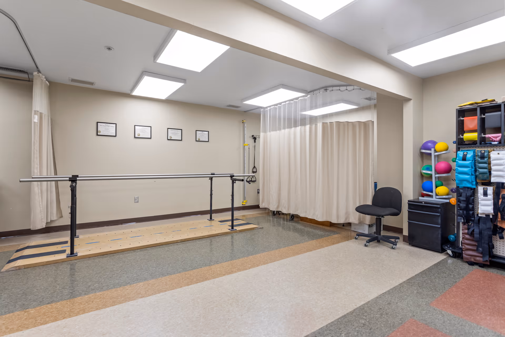 A physical therapy or rehabilitation room with parallel bars for walking exercises, a chair, a rack with colorful therapy balls and weights, and a curtained area in the corner. The room has a tiled floor and certificates framed on the wall.