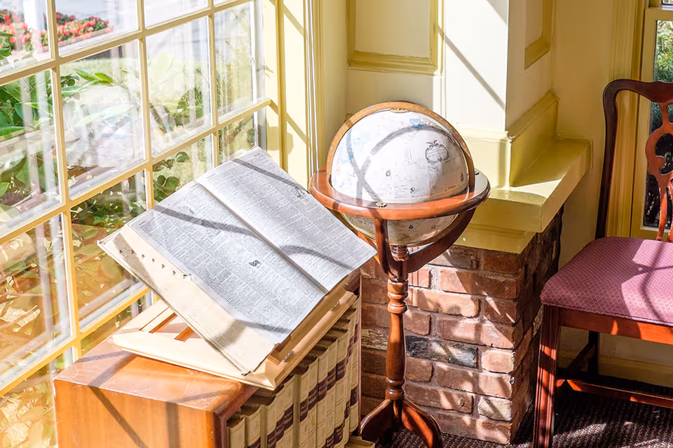 A sunlit corner of a room featuring a large open book on a wooden stand, a vintage globe on a wooden stand, and a wooden chair with a red cushioned seat. The corner has a brick base and cream-colored walls with decorative molding. A large window with multiple panes lets in natural light and shows some greenery outside.