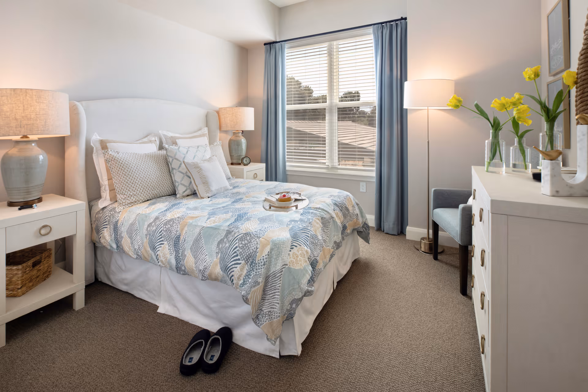 A cozy bedroom with a neatly made bed featuring a patterned quilt and multiple pillows. There are two bedside tables each with a lamp, a window with blue curtains letting in natural light, a floor lamp, a chair, and a white dresser with yellow flowers in vases on top. A pair of slippers is placed on the carpeted floor near the bed.