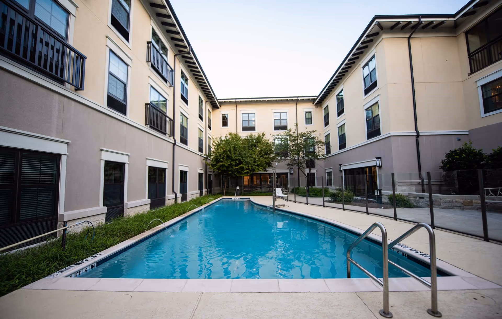 Outdoor swimming pool surrounded by a three-story senior living facility building with windows and balconies. The pool area is fenced with glass panels and has a metal handrail for entry. There are some trees and greenery near the building.