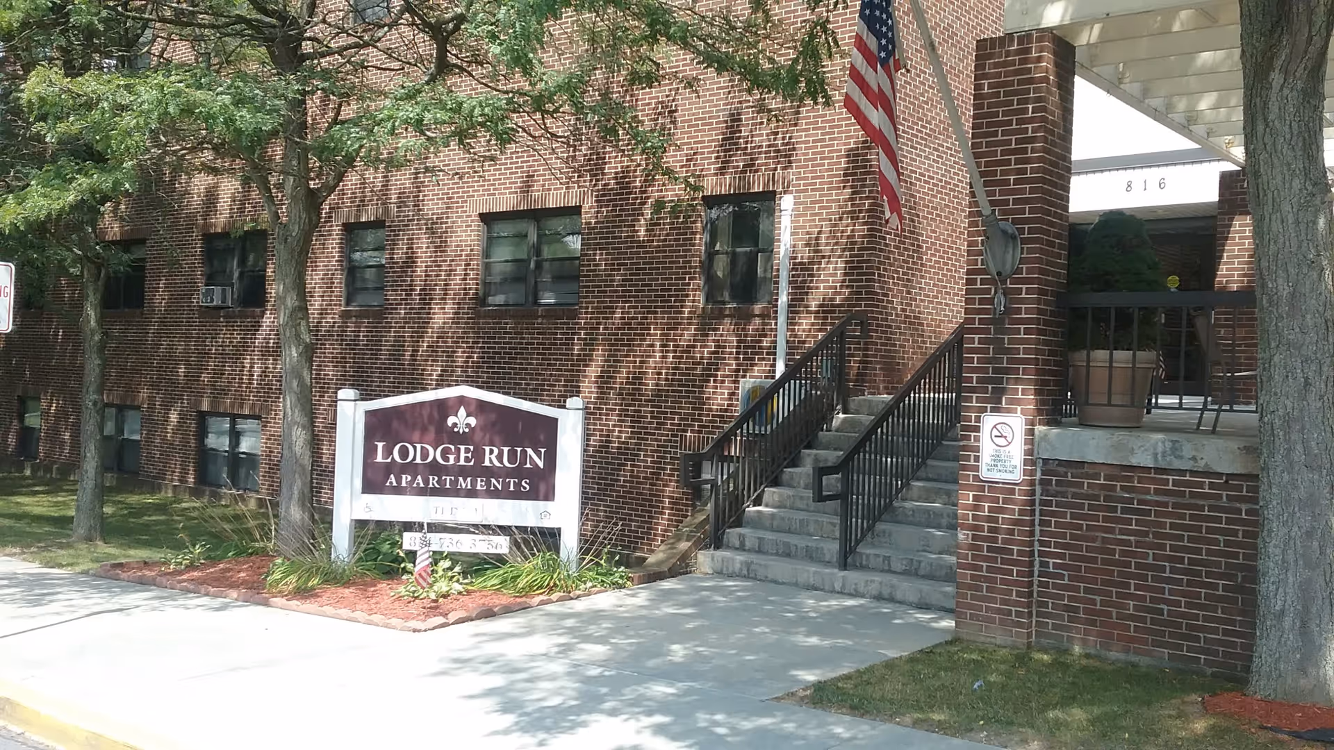 Exterior view of Lodge Run Apartments showing a brick building with several windows, a set of stairs with black railings leading to an entrance, an American flag mounted on the building, and a sign with the apartment name and contact number. Trees and landscaping are visible around the building.