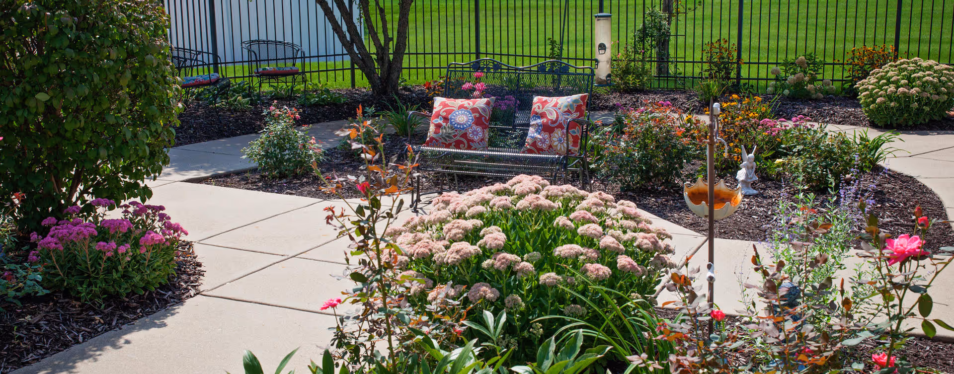 A well-maintained outdoor garden area with a paved walkway, various flowering plants, shrubs, and a black metal bench with two colorful patterned cushions. A black metal fence encloses the garden area.