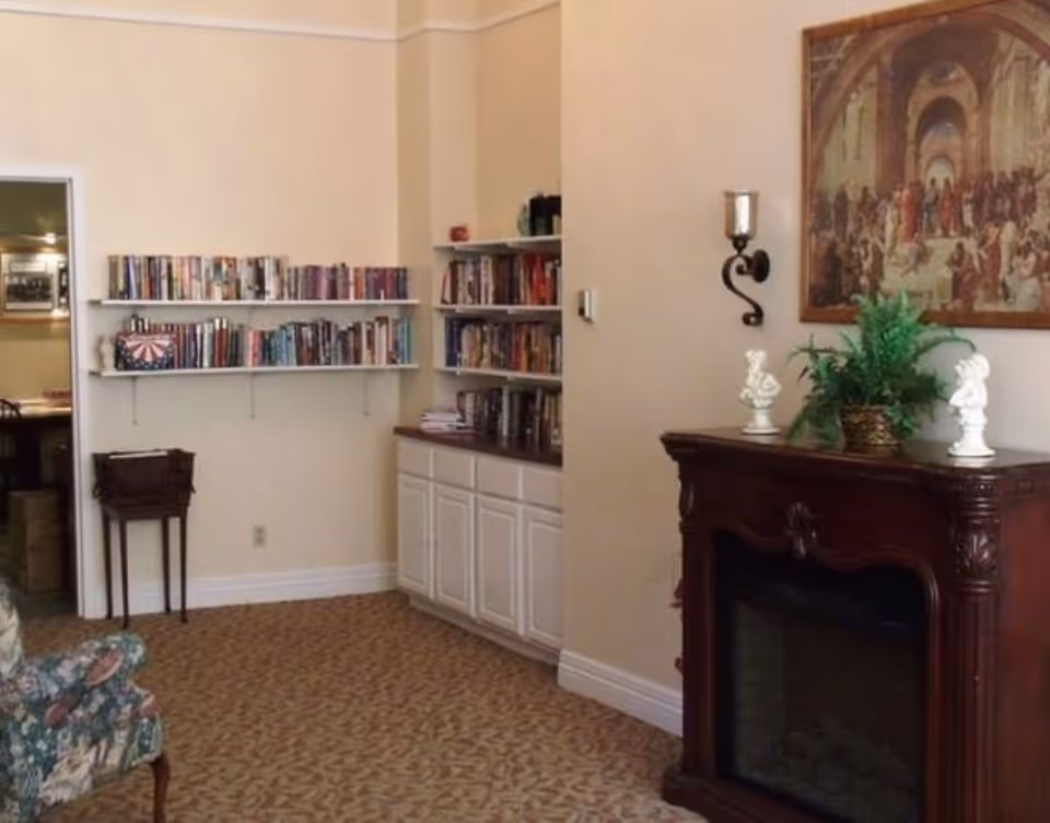 Interior room with beige walls and patterned carpet. There are white shelves filled with books on the left wall above a small wooden stand. A built-in cabinet with more books on top is in the corner. On the right side, there is a dark wooden fireplace mantel decorated with two white bust statues and a green potted plant. Above the mantel hangs a framed classical painting. A wall-mounted candle holder is also visible.