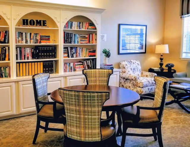 A cozy interior room featuring a round wooden table surrounded by four plaid upholstered chairs. Behind the table is a built-in white bookshelf filled with books and decorative items, including a sign that says 'HOME'. To the right, there is a floral armchair next to a side table with a lamp and a small plant. A framed picture hangs on the wall above the armchair, and a window with blinds allows natural light into the room.
