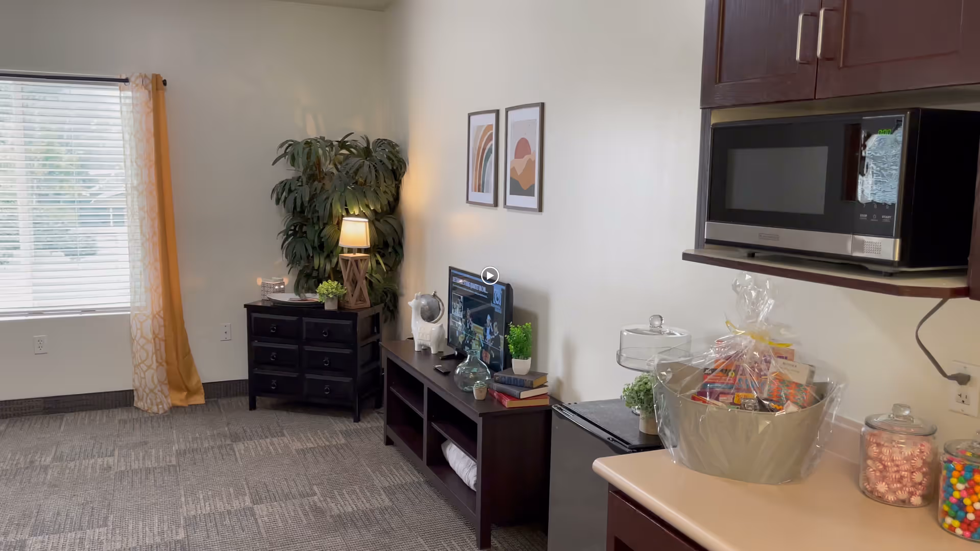 Sunlit assisted living sitting area with a TV stand, side table and lamp, potted plant, and a counter with a microwave, gift basket and jars of candy.