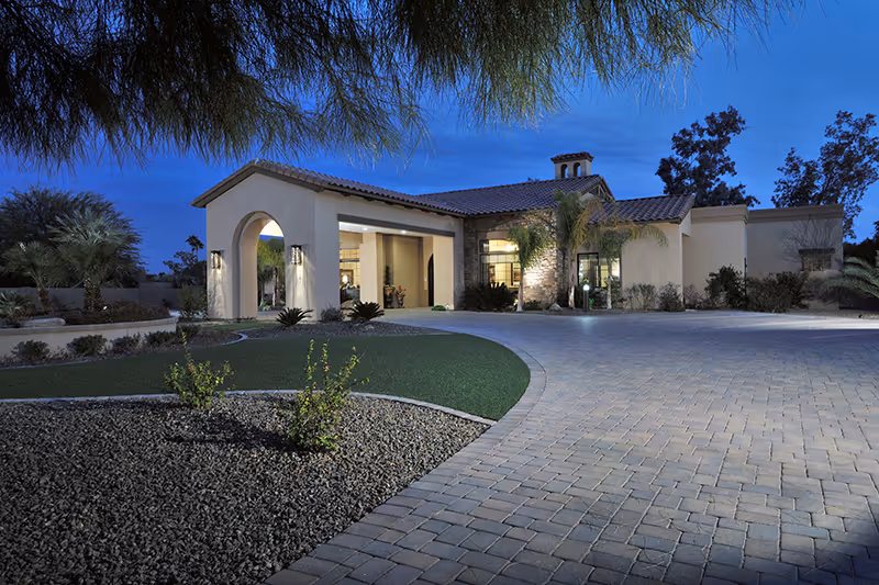 Front exterior of a single-story Mediterranean-style building with an arched entrance, paved driveway, and desert landscaping at dusk.