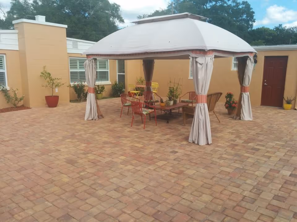 Outdoor patio area with a beige canopy tent covering a seating arrangement consisting of colorful chairs and a table. The patio is paved with brick tiles and surrounded by a tan building with windows and a red door. Several potted plants are placed around the patio.