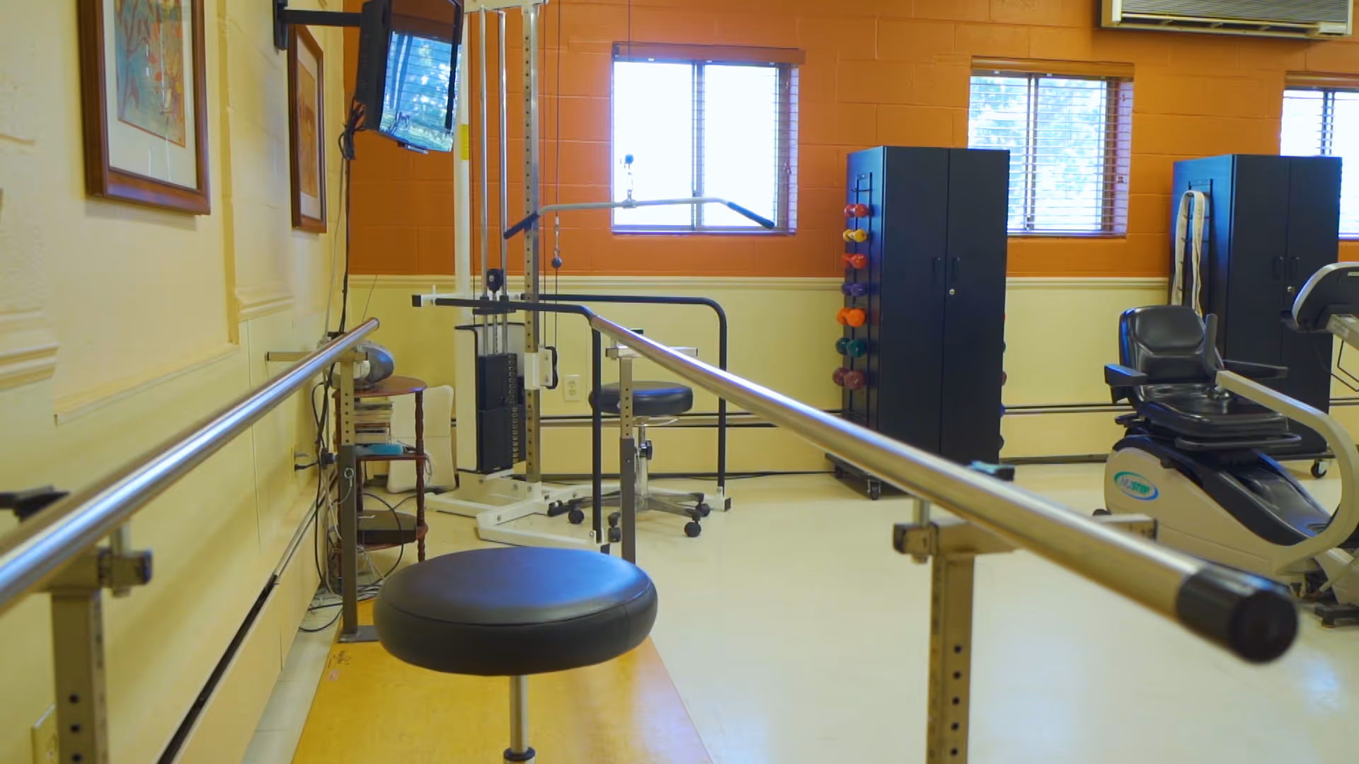 Physical therapy room with parallel bars for walking exercises, a padded stool, exercise equipment including a recumbent bike, weight racks, and two windows with blinds letting in natural light.