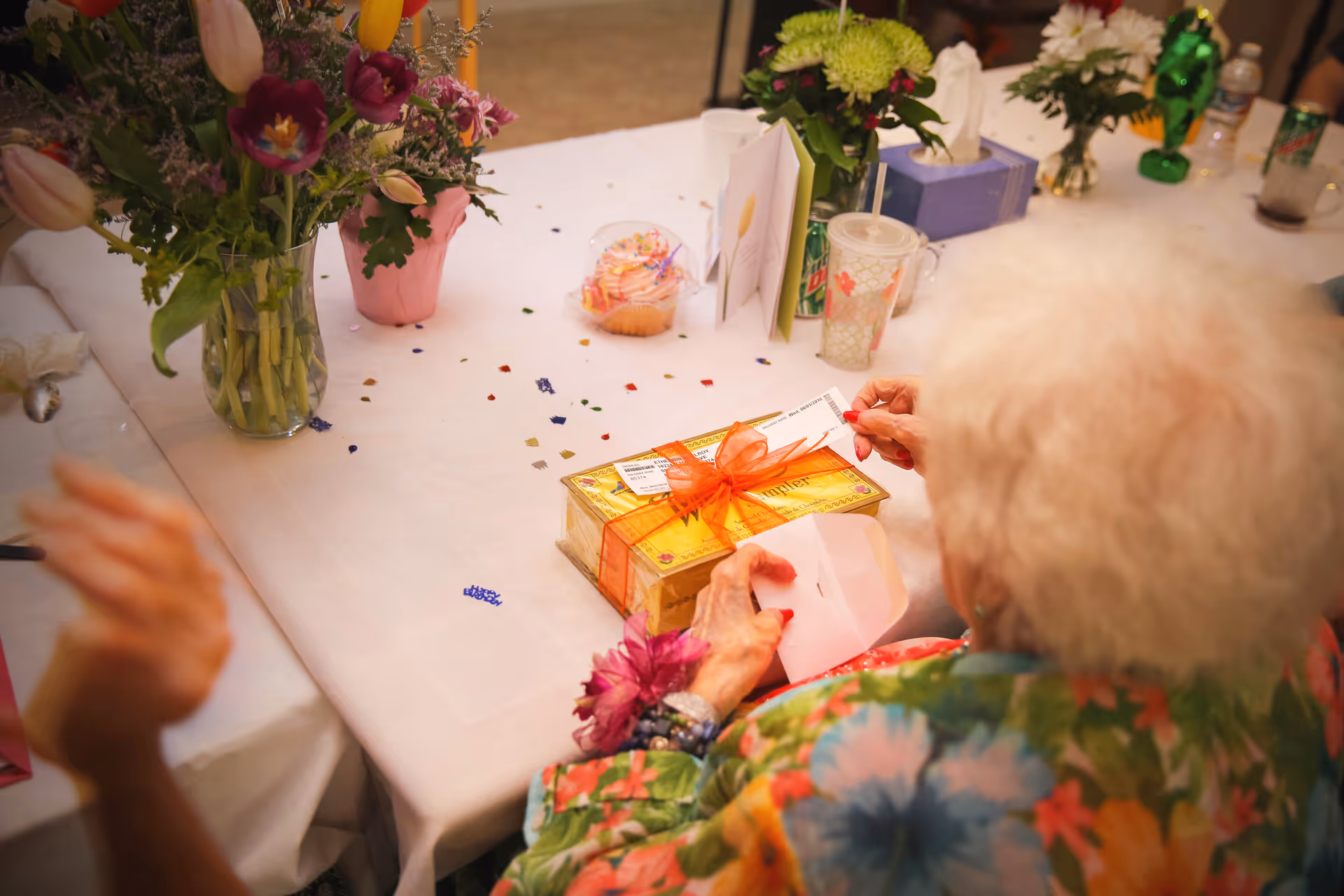 An elderly woman at a decorated table opens a wrapped gift box surrounded by flowers and party decorations.