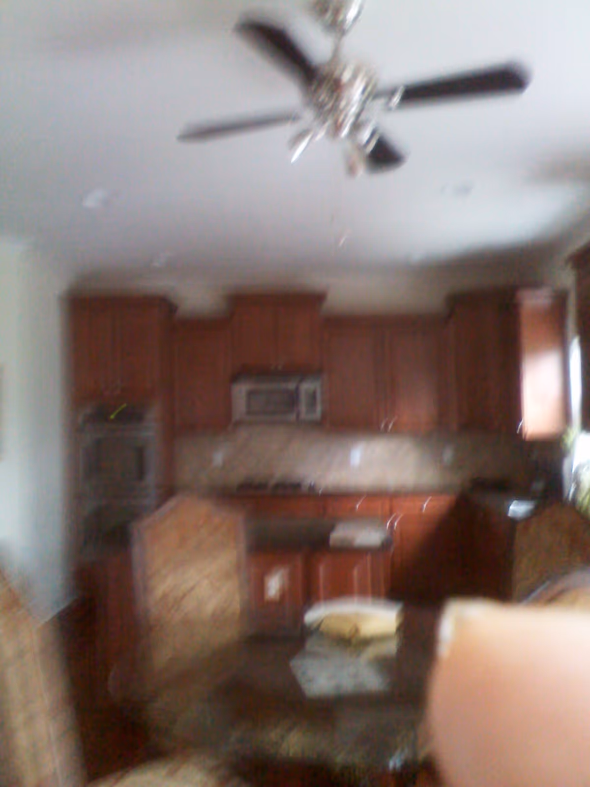 Blurry image of a kitchen with wooden cabinets, a ceiling fan, a microwave, an oven, and a dining table with chairs.