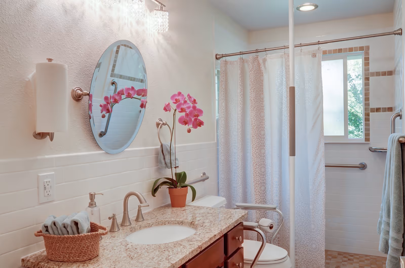 A bright and clean bathroom featuring a granite countertop with a sink, a round mirror above the sink, a pink orchid plant in a pot, a basket with rolled towels, and a soap dispenser. The bathroom has a shower area with a white patterned curtain, grab bars for accessibility, a window letting in natural light, and a towel hanging on a rack.