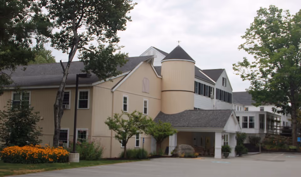 Front exterior of a beige multi-story senior living building with a covered entrance, turret-like tower, trees, and landscaped flowerbeds.