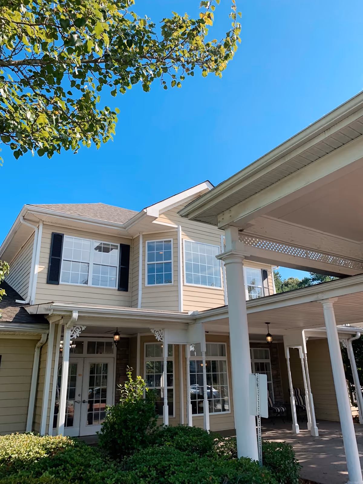 Exterior view of a two-story building with beige siding and white trim, featuring large windows and a covered entrance supported by white columns. Green bushes and a tree with green leaves are visible in the foreground under a clear blue sky.
