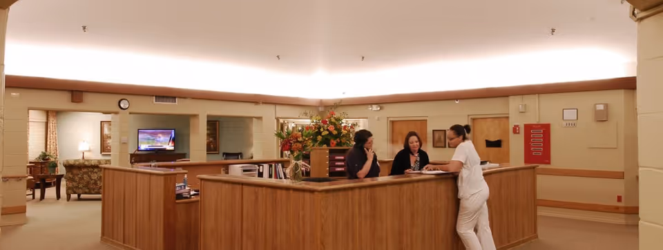 Wood-paneled reception desk in a senior living facility lobby with three staff talking and a visitor leaning on the counter and a seating area visible behind them.