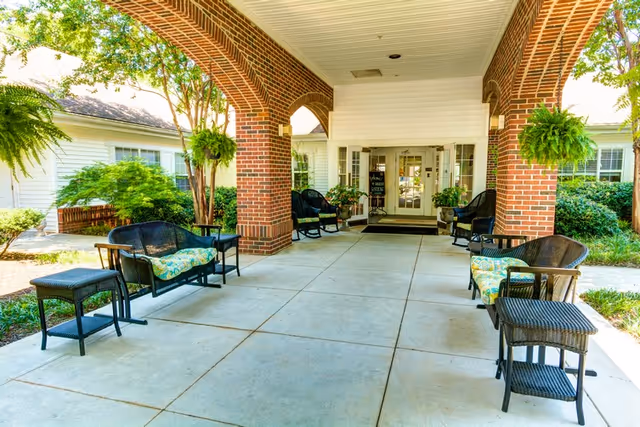 A welcoming entrance to Harbison Shores with a covered porch, featuring wicker chairs and potted plants, surrounded by greenery.