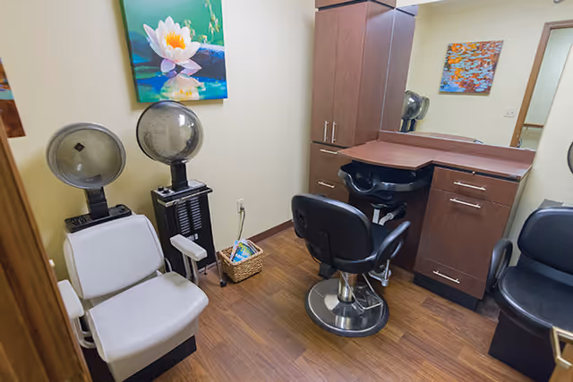 Small salon-style room with styling chairs, hooded hair dryers, a sink, cabinetry, and colorful wall art.