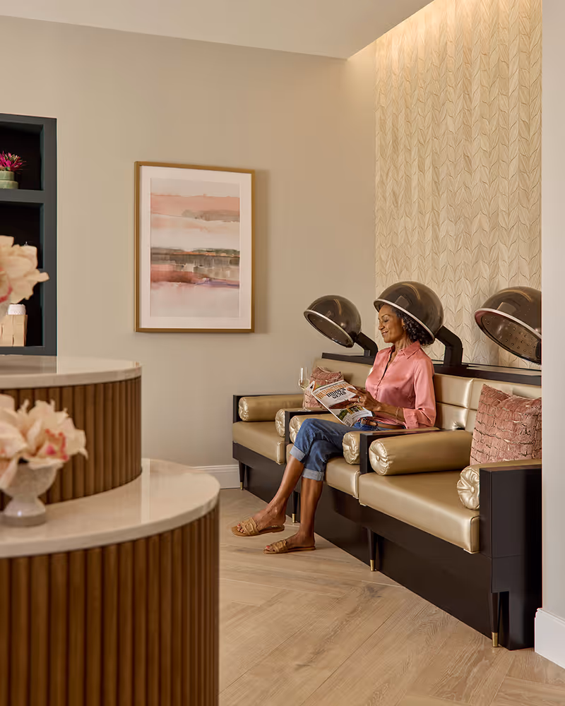A woman sits comfortably under a hair dryer in a salon area, reading a magazine and holding a glass of wine. The room features beige walls, a framed abstract painting, and a modern wooden counter with flowers in the foreground.